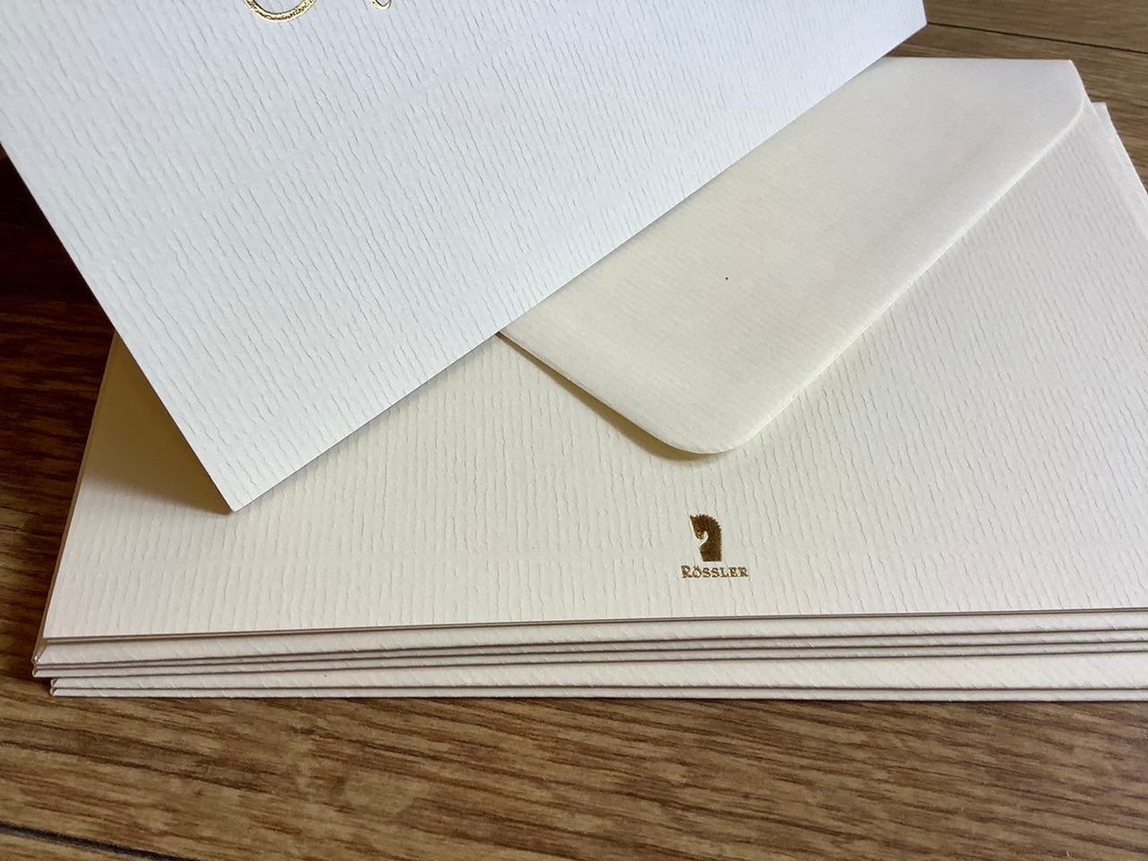 Edle Einladungskarten von Rössler Paper. Altweiße Klappkarte mit Umschlag, auf der Karte steht in eleganter Schreibschrift Einladung (Goldfolienprägung). Geripptes Papier.