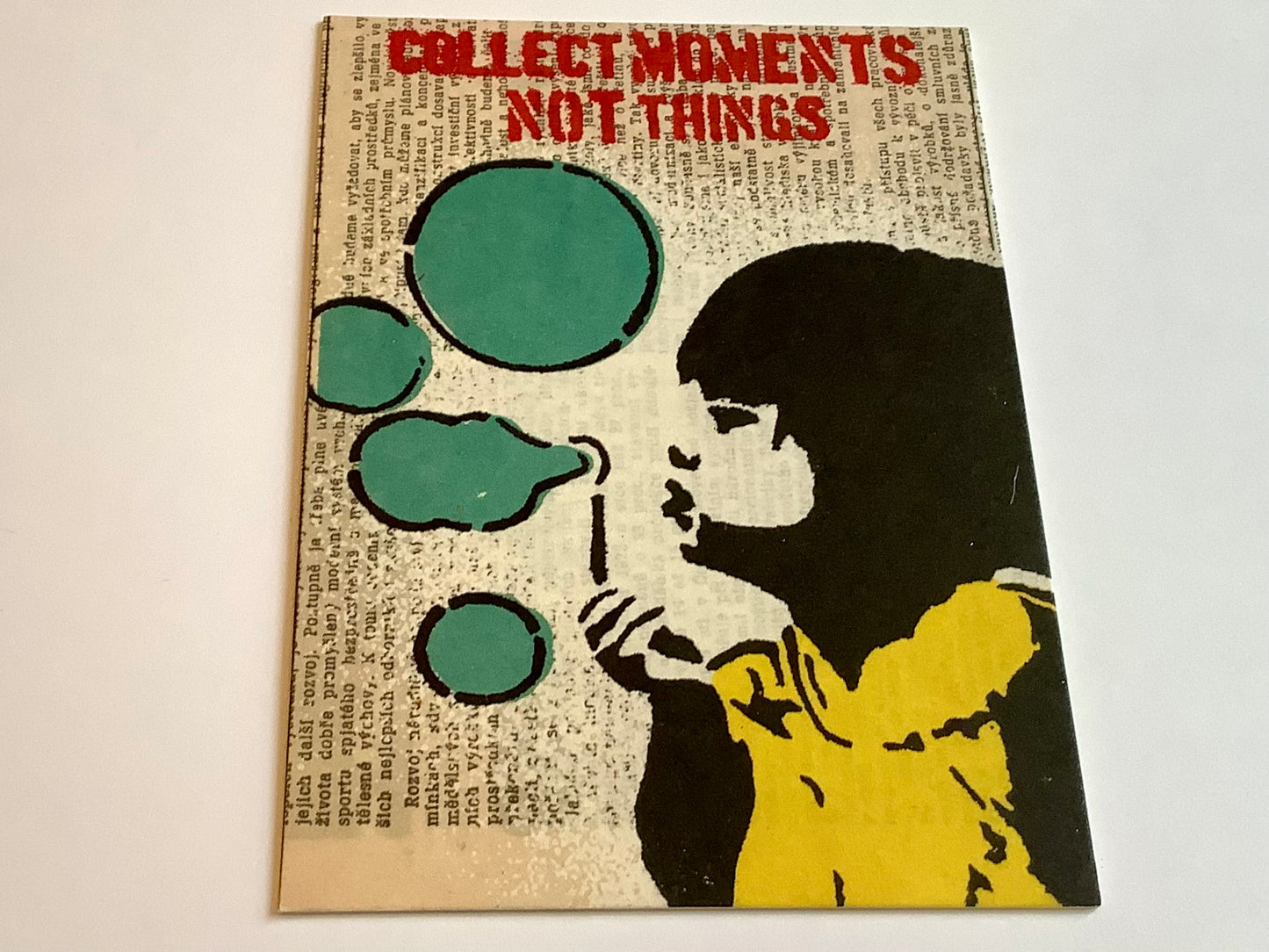 Auf der illustriertern Postkarte sieht man ein Mädchen mit Seifenblasen im Graffiti-Schablonenstil von Banksy. Darüber der Satz "Collect moments not things"
