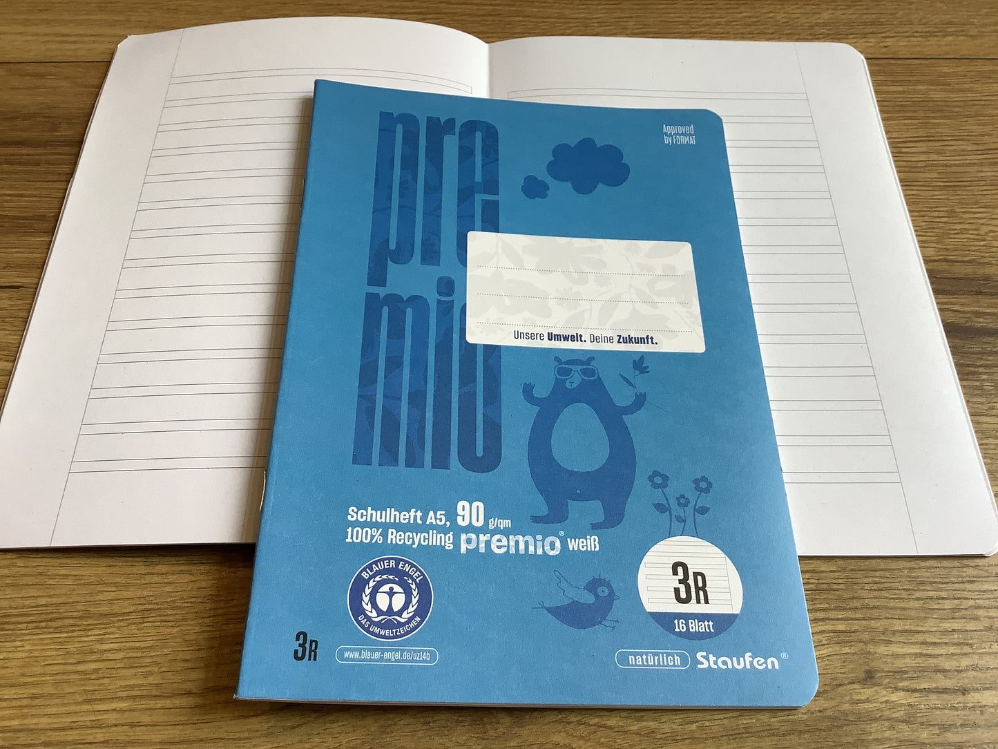 kleines Schulheft mit blauen Cover, einmal geschlossen, darunter einmal aufgeschlagen mit den „Schlauch-Linien“ fürs dritte Schuljahr