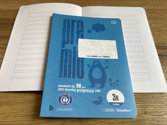 kleines Schulheft mit blauen Cover, einmal geschlossen, darunter einmal aufgeschlagen mit den „Schlauch-Linien“ fürs dritte Schuljahr