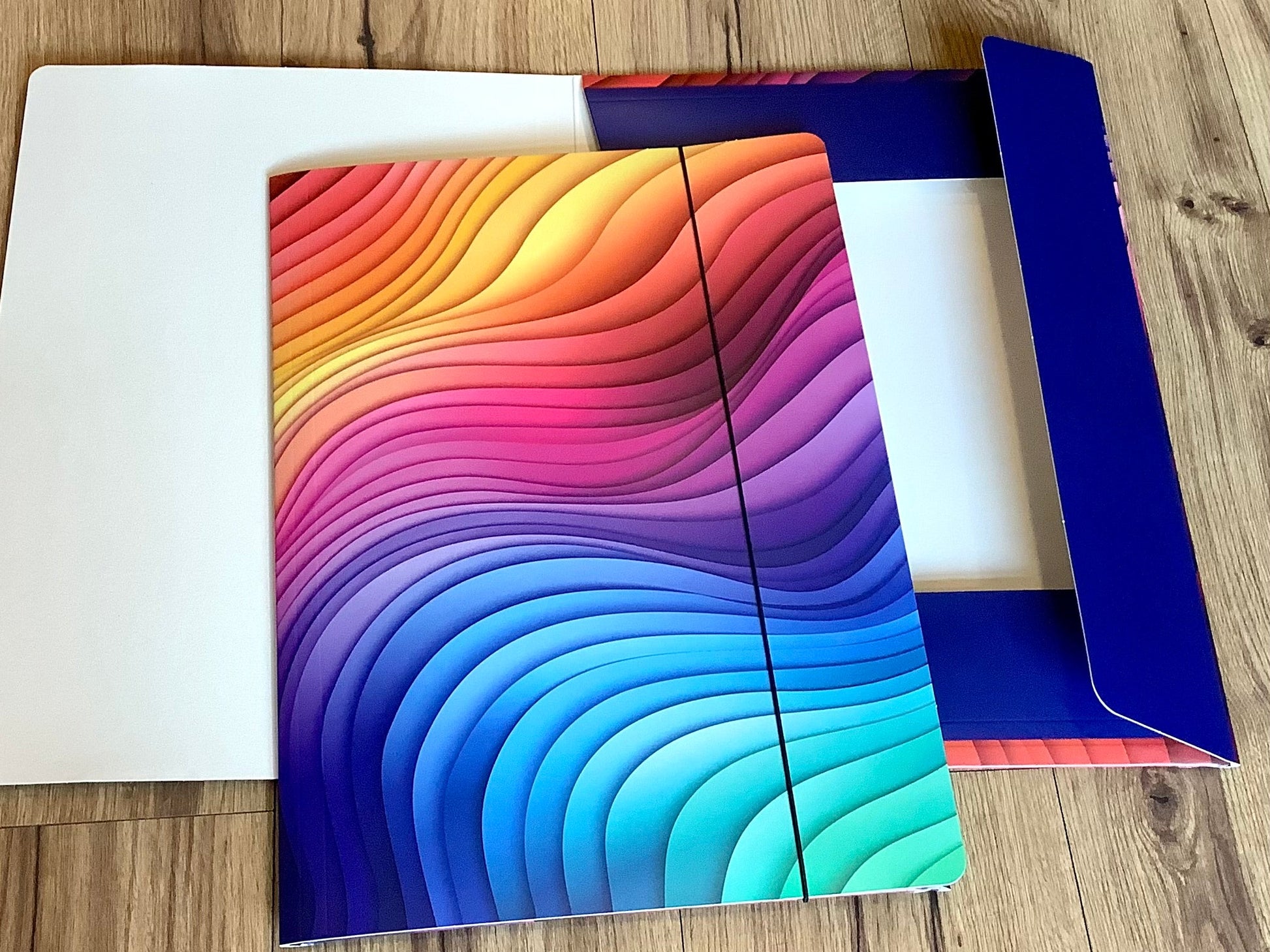 In grafischen Wellen in Regenbogenfarben bedruckte Sammelmappe mit Gummizug aus Recyclingpappe. 
Erhältlich  bei Polly Paper - umweltfreundlicher Schulbedarf in Berlin-Mitte