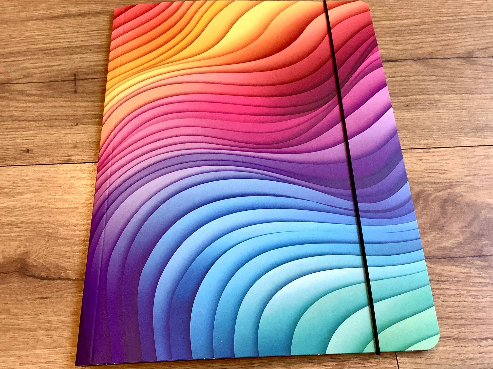 In grafischen Wellen in Regenbogenfarben bedruckte Sammelmappe mit Gummizug aus Recyclingpappe. 
Erhältlich  bei Polly Paper - umweltfreundlicher Schulbedarf in Berlin-Mitte