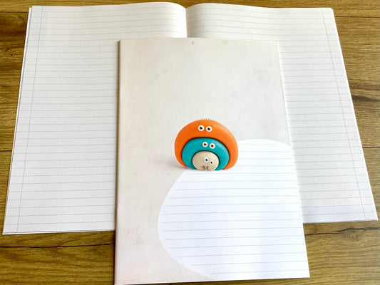 Ein liniertes Heft, das nicht nach Schule aussieht: süße bunte Knetfigur (Fantasiefigur) auf dem Cover. Weiß, liniert mit schmalem Rand, durch eine Linie abgetrennt.